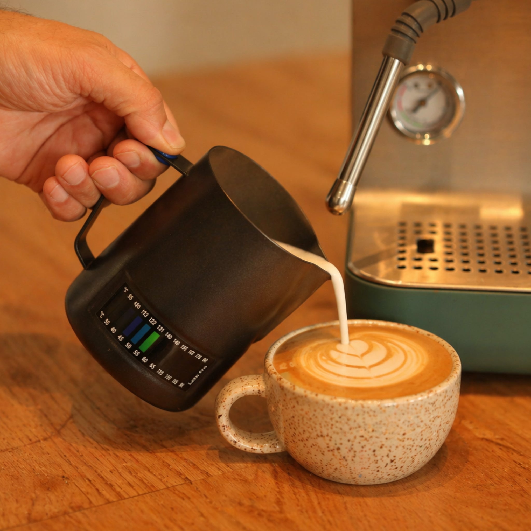 Person pouring coffee from a black thermometer milk jug into a cup with latte art on a wooden surface.
