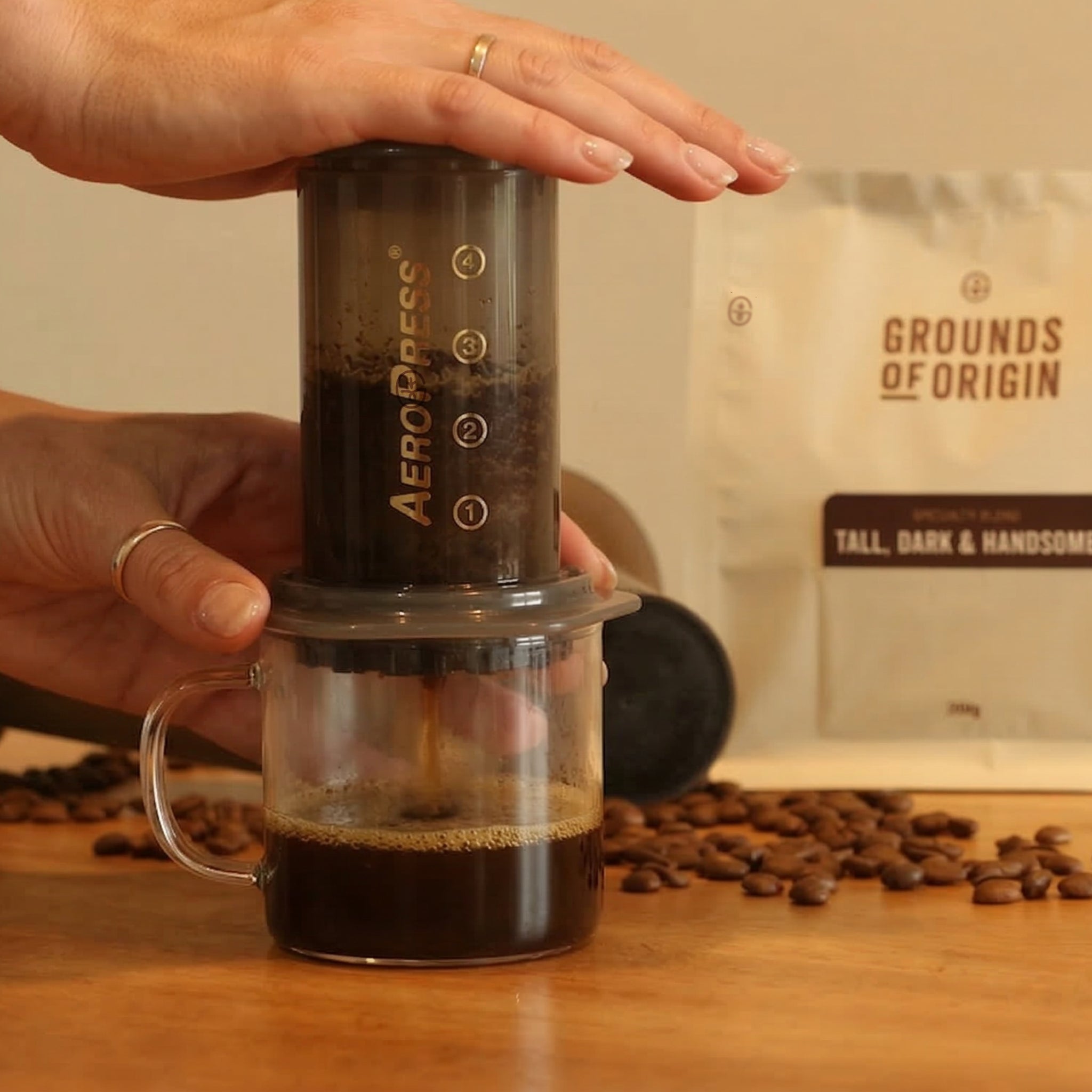 A person using an Aeropress to make coffee with a bag of Grounds of Origin coffee beans in the background.