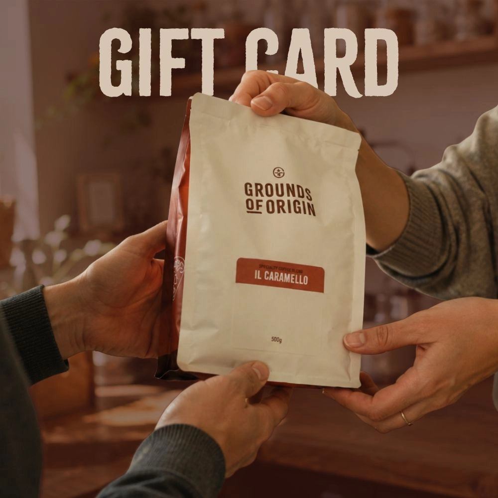 Two people gifting a 'Grounds of Origin' coffee bag with 'Il Caramello' flavor, labeled as a gift card.