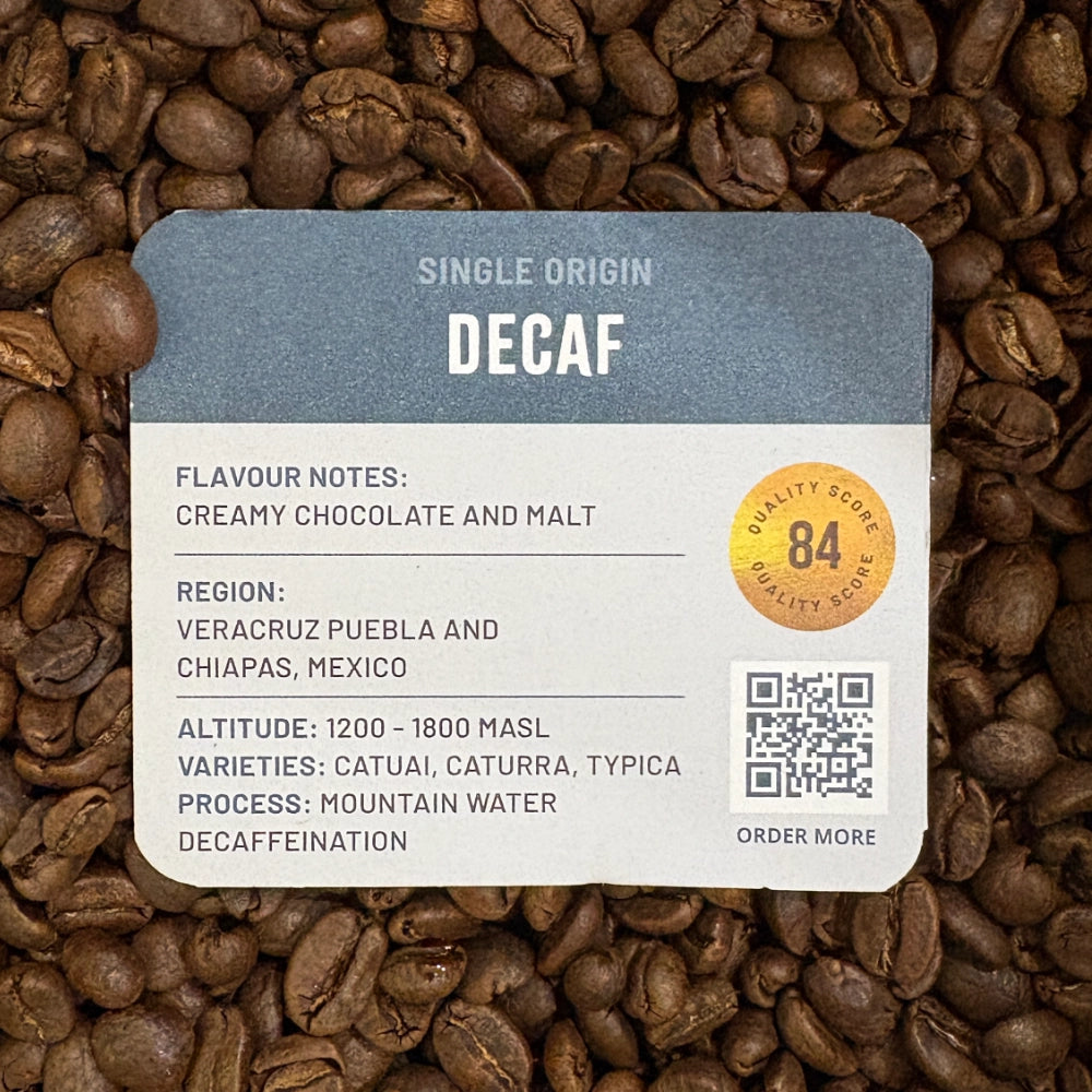 A close-up of Grounds of Origin coffee beans with a printed label reading 'Single Origin Decaf' and flavor notes, placed over a bed of roasted coffee beans. The label is slate blue on top and white on the bottom, with a gold quality score badge and a QR code.