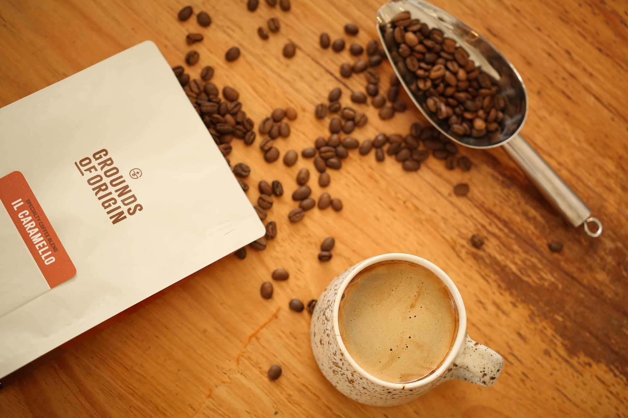 Coffee cup, coffee beans, and a scoop on a wooden surface with 'Grounds of Origin' packaging.