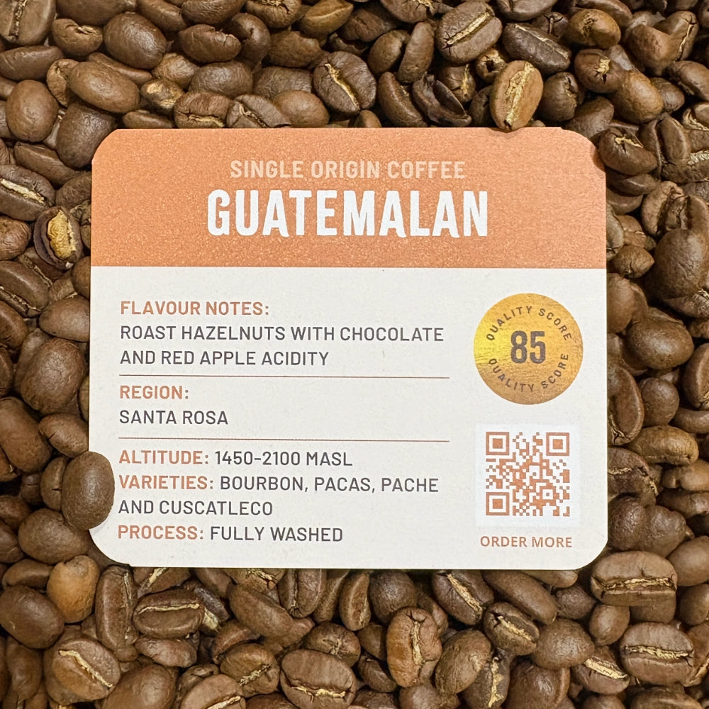 A Grounds of Origin coffee label card placed over a close-up background of roasted coffee beans. The label reads 'Single Origin Coffee Guatemalan' with flavor notes, region, altitude, varieties, process, and a quality score of 85.