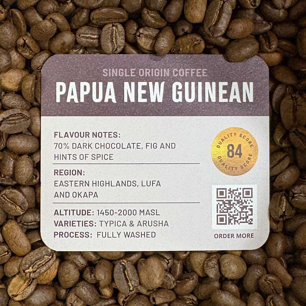 A Grounds of Origin coffee label reading 'Single Origin Coffee Papua New Guinean' placed over a background of roasted coffee beans. The label includes flavor notes, region, altitude, variety, processing, a quality score badge, and a QR code.