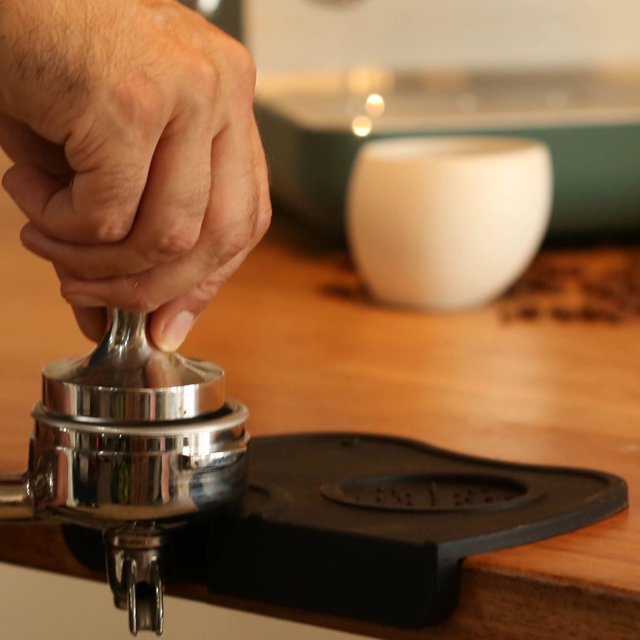 Hand using a metal espresso tamper on a black mat with a blurred cup in the background.