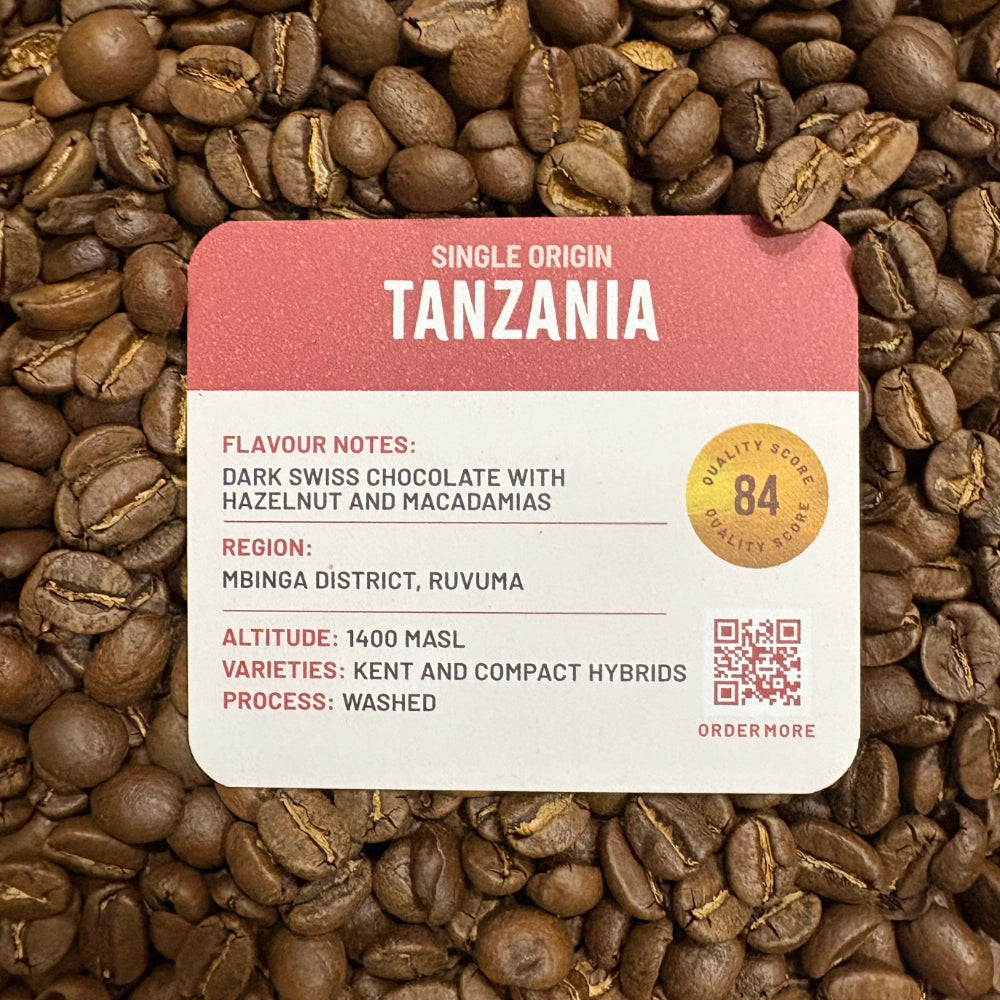A Grounds of Origin coffee label card placed over a dense background of roasted coffee beans. The label reads 'Single Origin Tanzania' with flavor notes, region, altitude, varieties, process, a quality score badge, and a QR code.