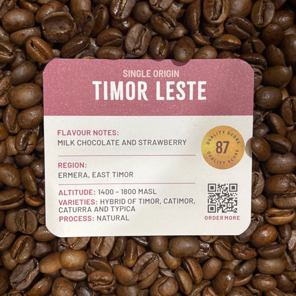 A Grounds of Origin coffee label for Timor Leste single origin coffee, placed over a background of roasted coffee beans. The label has a dusty pink top section with white text, and a cream-colored bottom section with black text detailing flavor notes, region, altitude, and variety.